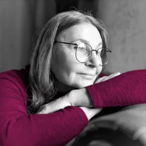 Eine Frau mittleren Alters mit Brille sitzt auf einer Couch und guckt gedankenverloren nach draußen.